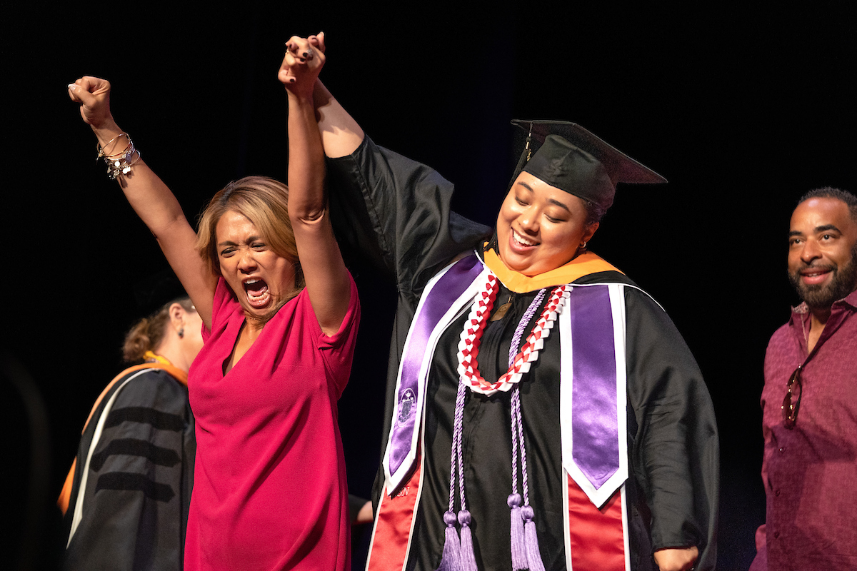 This is a photo of a group of four people at a graduation ceremony. One person is wearing a graduation cap and gown, and another is wearing a pink dress and has their arms raised in celebration. The background is a stage with a black curtain.