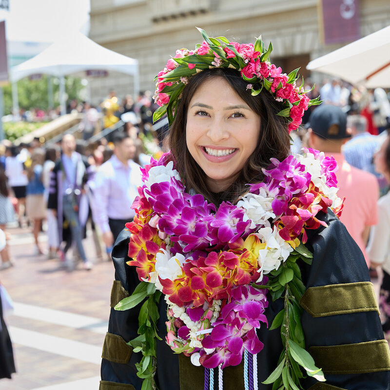 Pictured here is one graduate covered in bright and beautiful flower leis. They are also wearing a crown of flowers on their head. In the background are several family members and graduates gathered together in celebration.