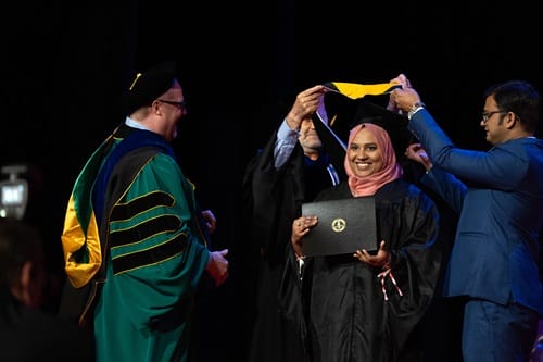 This images shows a graduate smiling at the camera while her family hooders place her hood over her shoulders.