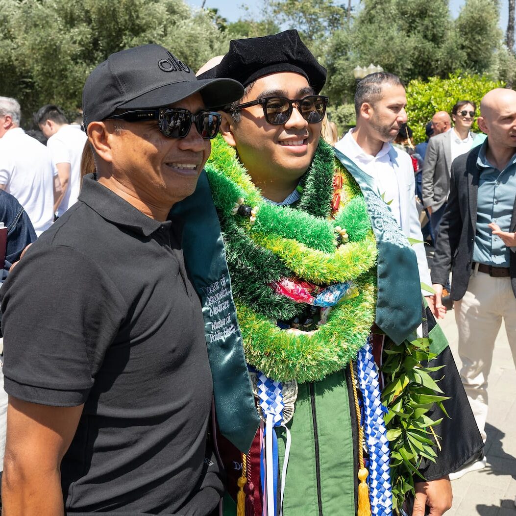 A graduate wearing a green academic gown, doctoral cap, and multiple colorful leis smiles while posing for a photo with a supporter outside the commencement ceremony. The supporter is wearing a black polo shirt, sunglasses, and a cap. Other attendees and greenery are visible in the background.