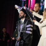 051325 WesternU CDM Commencement 115 A smiling male graduate in a black gown and cap holds his diploma case on stage. Two academic officials, a man and a woman, are placing a purple and white academic hood over the graduate's head during the hooding ceremony.