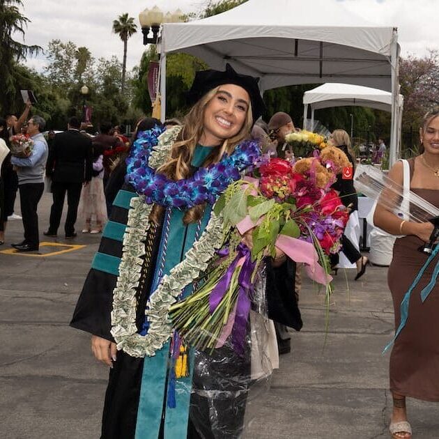 A smiling graduate in a black and teal academic gown and velvet cap stands outdoors, holding a large bouquet of flowers and wearing multiple colorful leis, including one made of currency. She is posing with a group of three younger women who are also smiling, in a sunny outdoor plaza with palm trees and white tents.