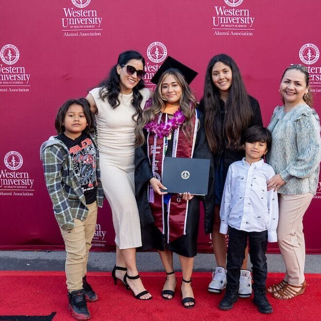 A graduate in a black cap and gown, maroon stole, and flower leis stands on a red carpet with five family members (two women and three young boys). They are posing in front of a tall, red backdrop with the Western University Alumni Association logo and a vertical banner to the right that reads "CONGRATULATIONS GRADUATES."