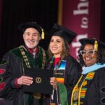 051425 CHS Commencement 084 A smiling female graduate in a black cap and gown, wearing a striped serape-style stole, stands between President Farias-Eisner and Dean Evans Grayson. All three are smiling and the graduate is holding a diploma folder. The officials are in black doctoral regalia, and the background is a dark stage with maroon banners