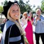 25VET Grad A smiling female graduate with long blonde hair and a black and gray academic gown and cap poses for a photo outdoors. In the background, three people are taking pictures of her with their phones.