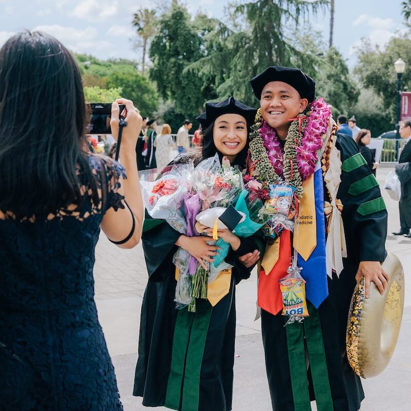 Two graduates in black gowns with green velvet panels and doctoral caps stand outdoors smiling and holding bouquets. They are covered in colorful leis and ribbons. A woman in a navy lace dress stands in front of them taking their photo. Trees and people are visible in the background on a sunny day.