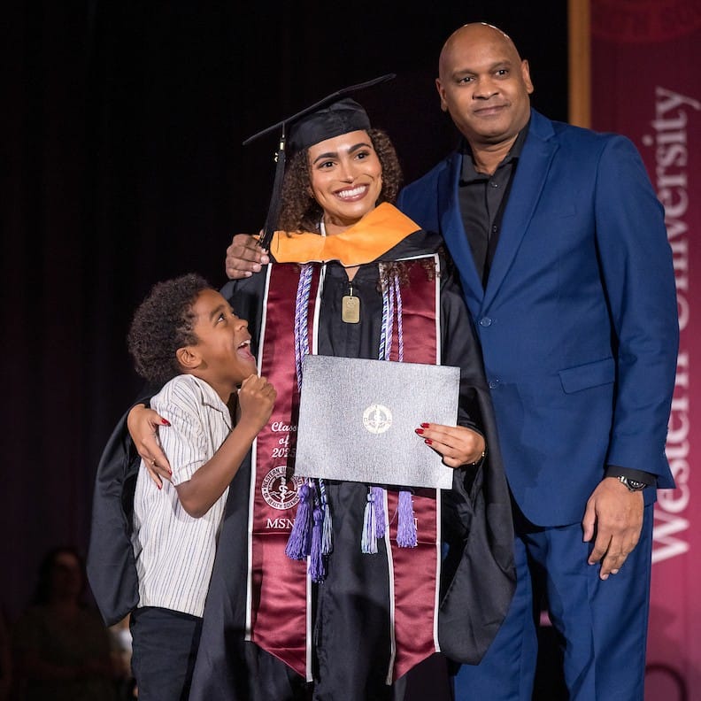Image of a graduate posing with a man in a blue suit and a child wearing a white shirt. The child is looking up and excitedly smiling at the graduate.